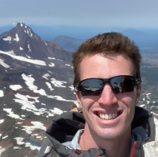 A scientist stands on a bare, rocky slope of a mountain, punching a steel tube into the ground with a glass sample bottle on top