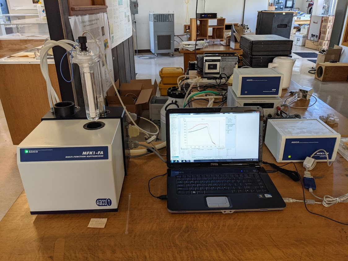 A blue-and-beige instrument box with a glass tube suspended like a streetlamp above it sits on a table next to a laptop computer