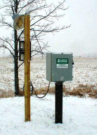 Equipment box with USGS label on top of iron pipe in ground with satellite reciver on pole next to it in snow covered field