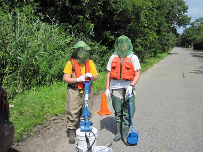 2 women on rural blacktop with safety vests and mosquito netting on heads with sediment sampling equipment