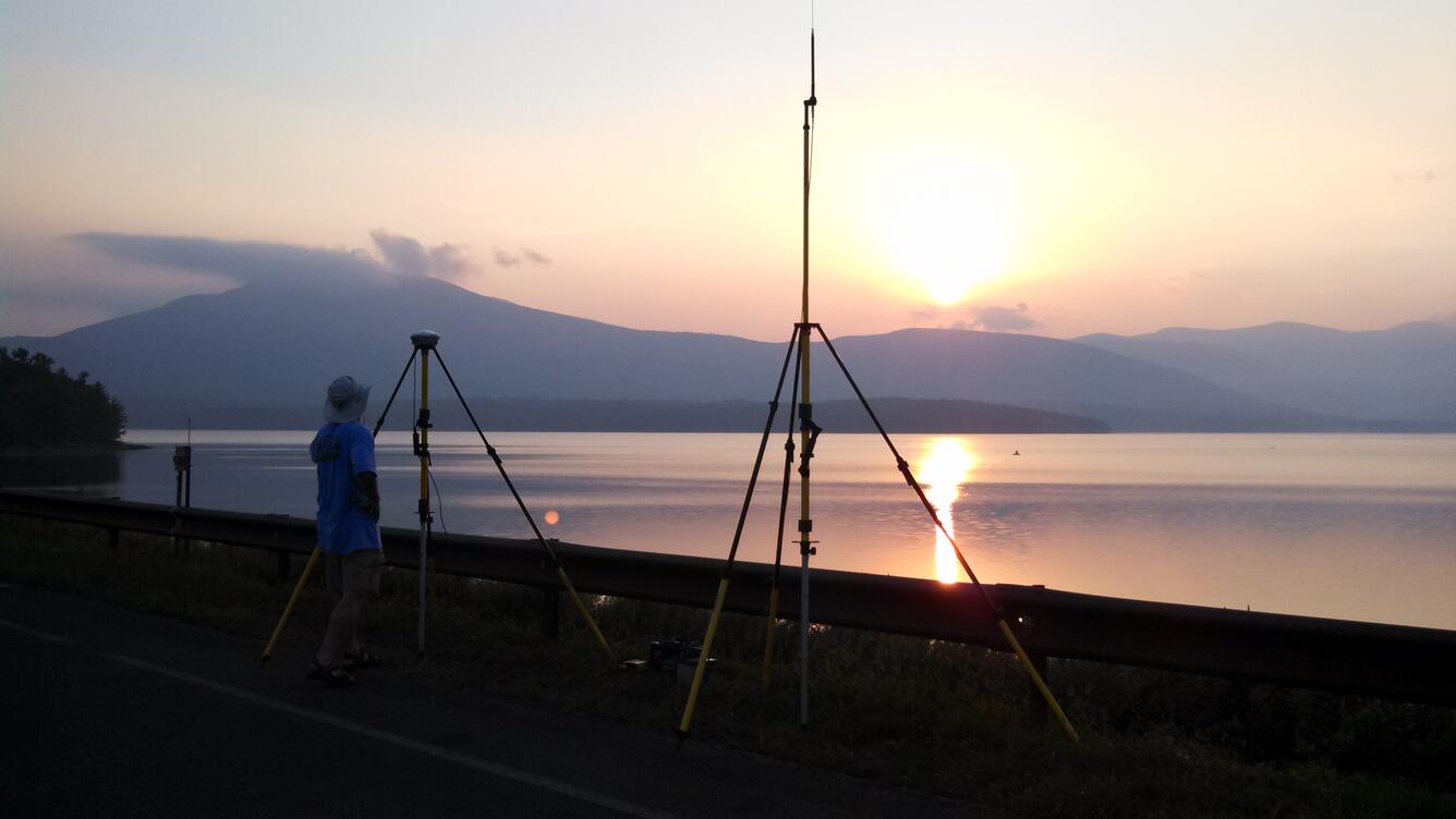 Man standing on shore by GPS base station with sunset over the reservoir and mountains