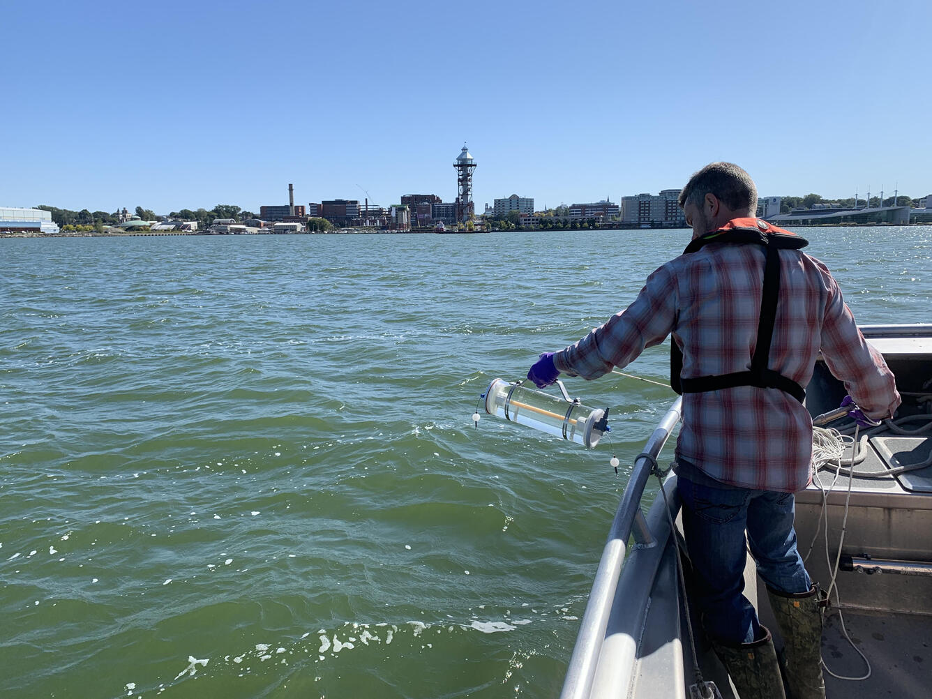water, sky, city, buildings, man, plaid, boat, trees, skyline