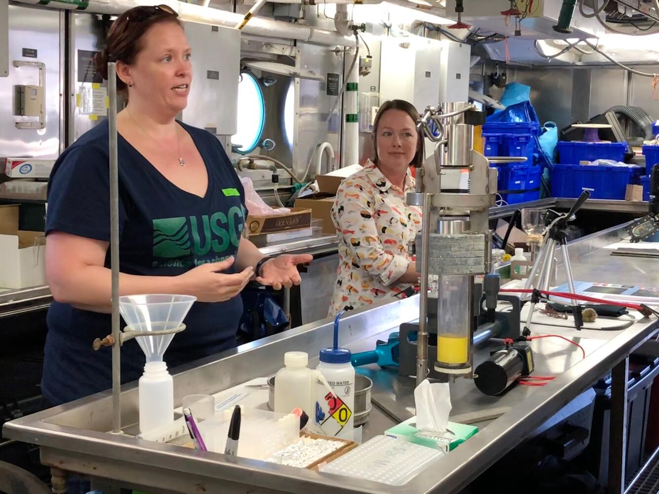 A woman stands behind a lab table talking and gesturing with her hands, while another woman stands off to her side.