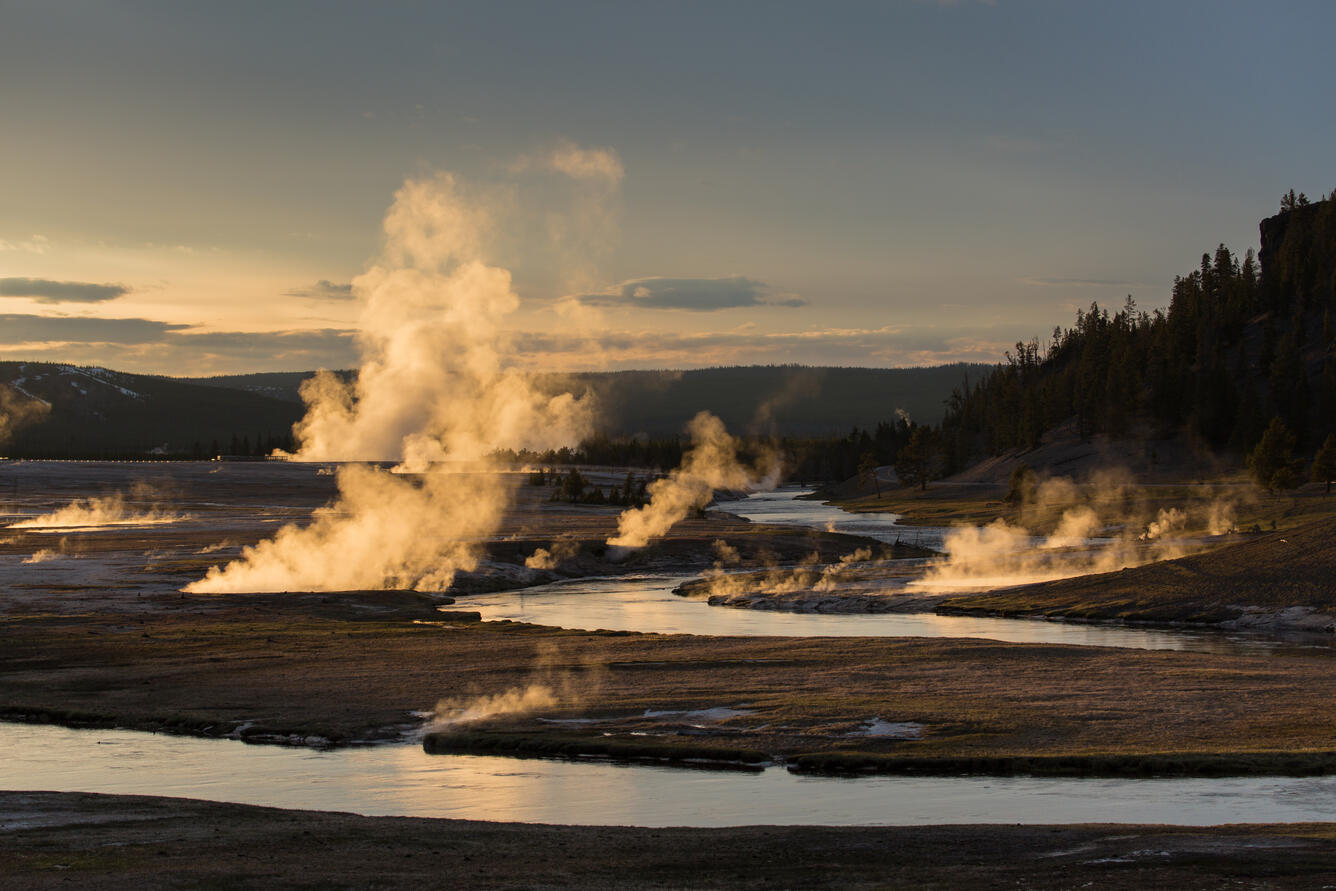 river meandering through wide valley, with wisps of stem rising at various spots. Illuminated at sunset, with golden reflections