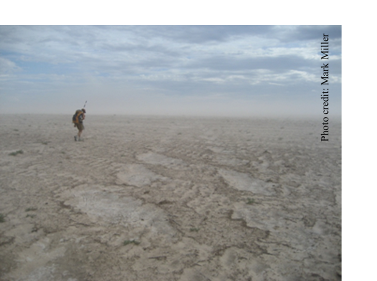 A lone researcher walking across a dusty, dryland landscape devoid of plants