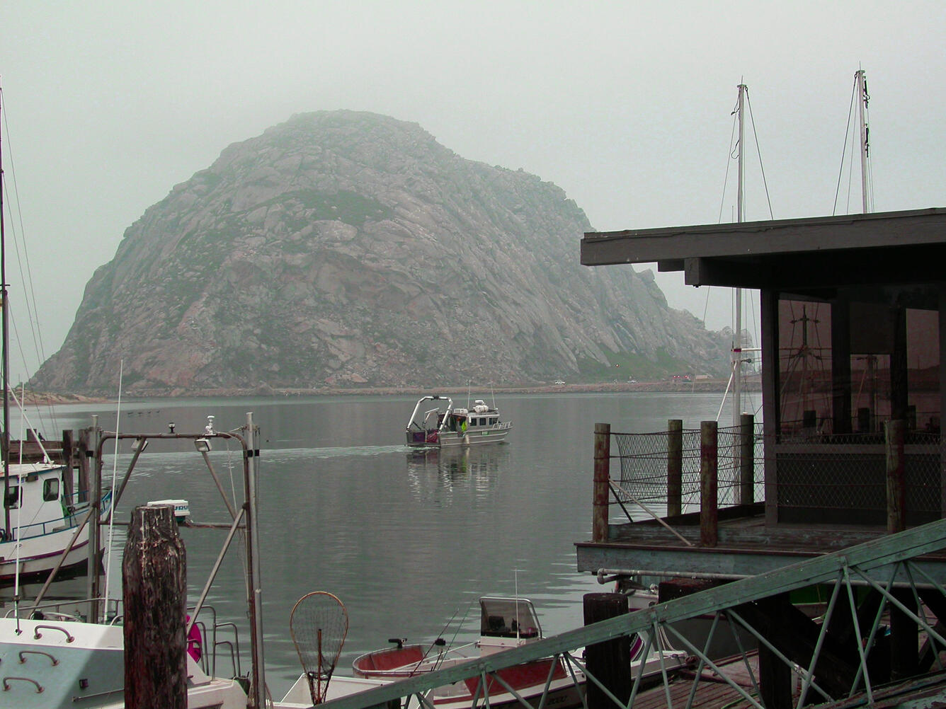 View from a dock near a calm bay with boats docked and one motoring slowly through the bay with a large outcrop in background.