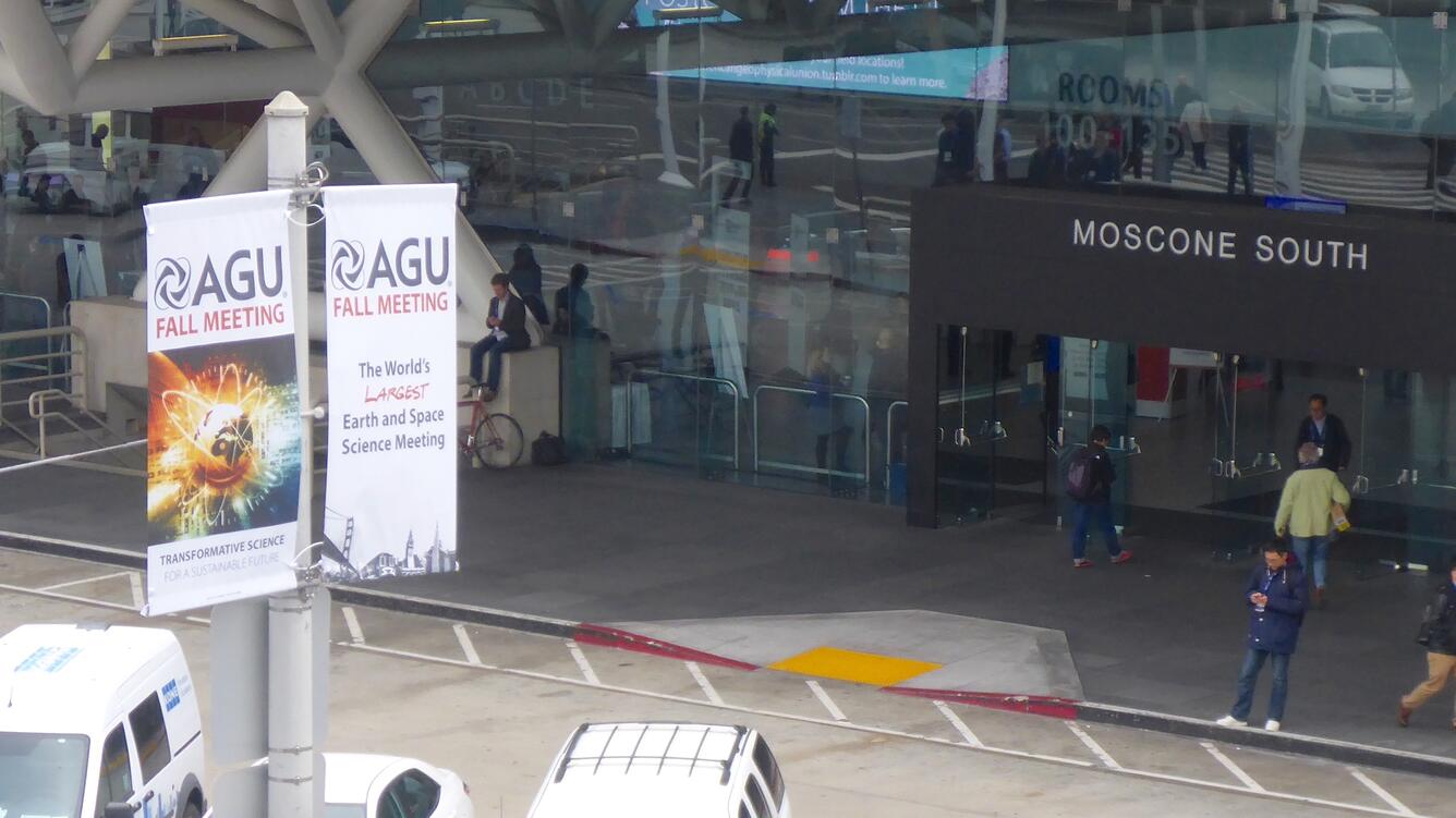 View looks at the entrance to a glass-front building with some cars parked nearby and some pedestrians walking on sidewalk.