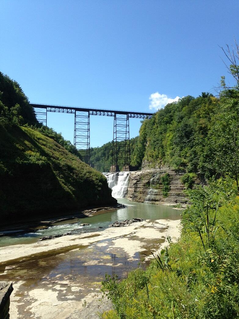 tall bridge over waterfalls flowing over rockface into a river with forest hills on both sides