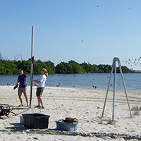 Scientists operate scientific sediment coring equipment with a tall metal barrel and tripod on a sandy beach near the water.