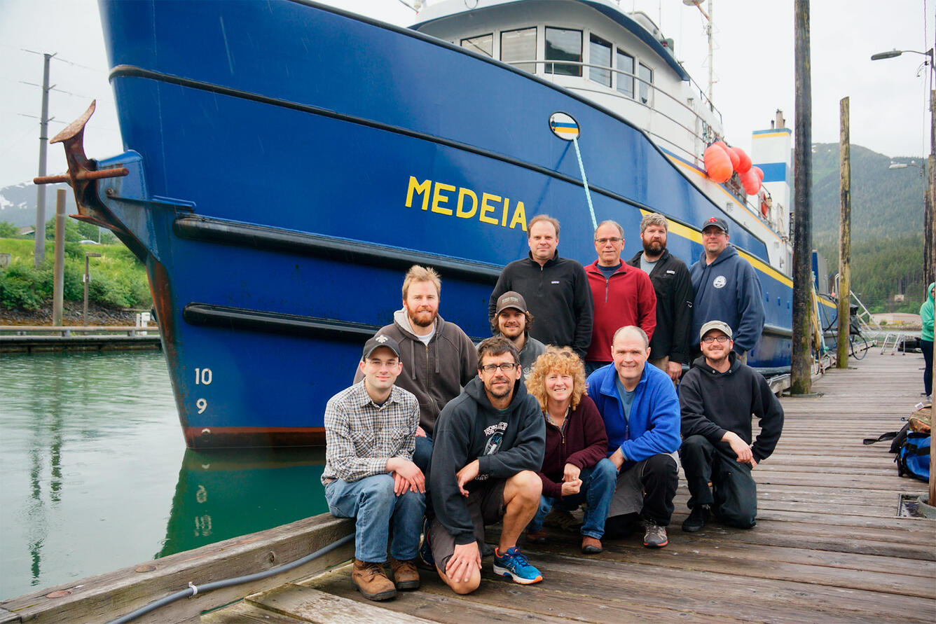 A group of scientists pose on the dock in front of a ship with a blue hull.