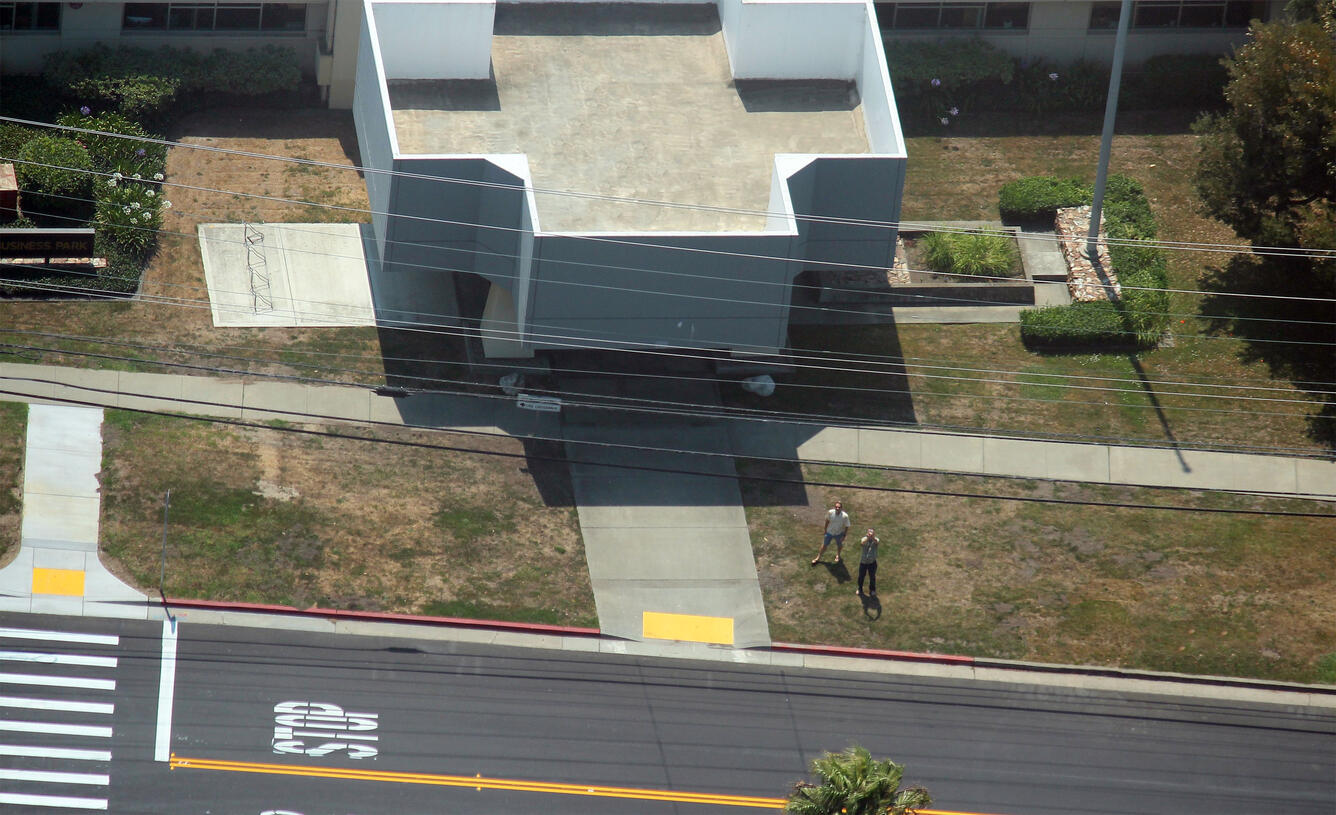 View from the sky looking down on a roadway with a crosswalk, some grass and trees, a building, and two men looking up.