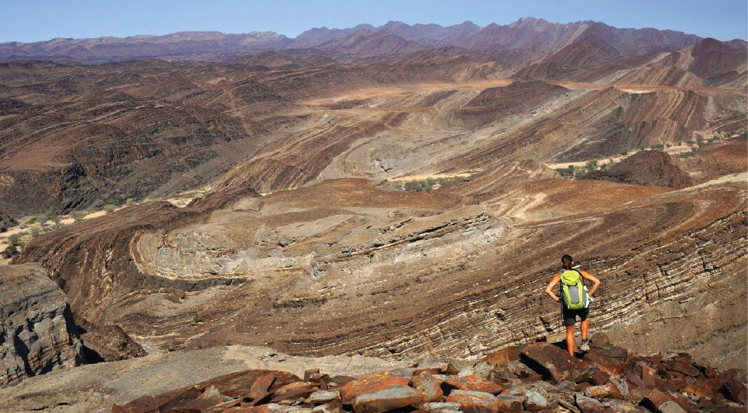 A person wearing a backpack walks across rocks in a rocky hilly area.