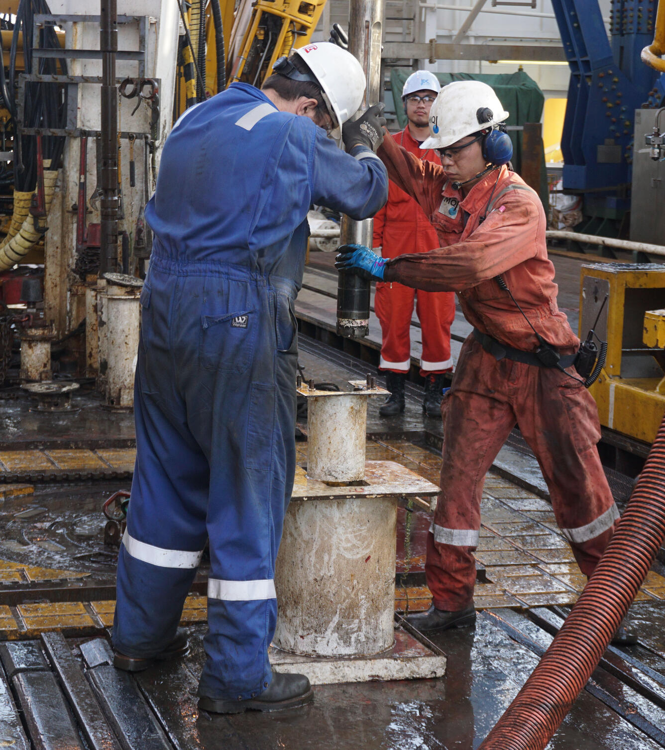 Image shows two scientists in hard hats collecting a research core aboard a drill rig