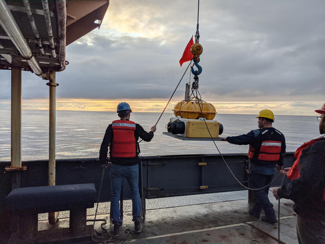 Two people on a ship guide an object, bolted to a metal platform with a plastic housing, over the side of the ship.