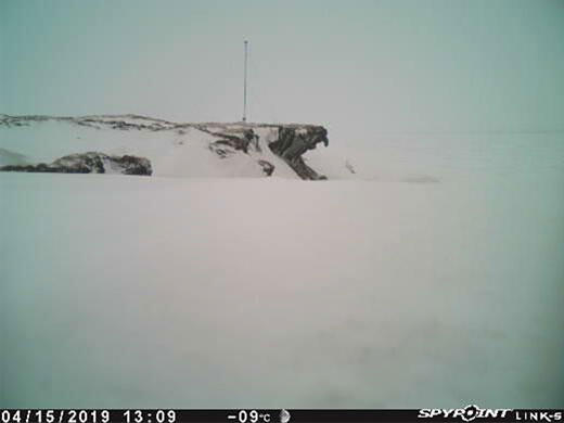 A ground-level view filled mostly with snow, with an arctic bluff in the background that has a pole mounted on it.