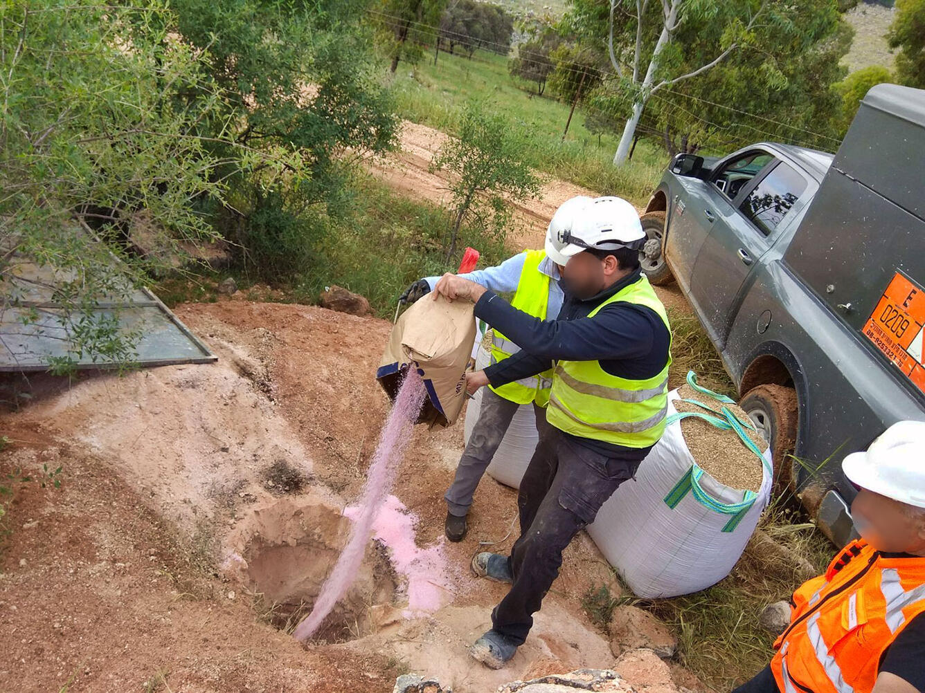 Workers wearing safety gear and hard hats pour pink powder into a hole in the dirt. another person looks on.