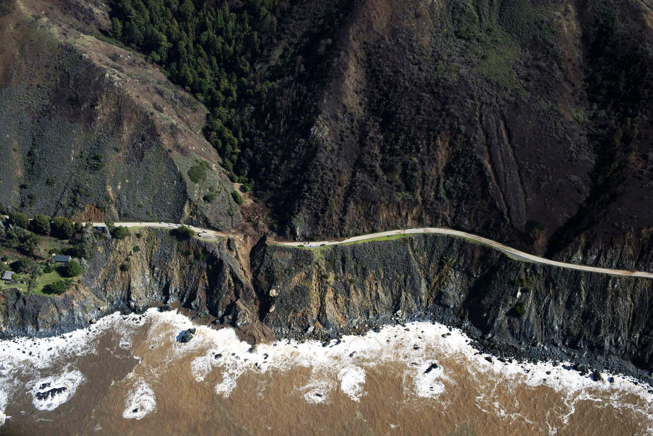 A view from the sky of a winding coastal road that runs along steep, rocky cliffs, with a section washed out and collapsed.