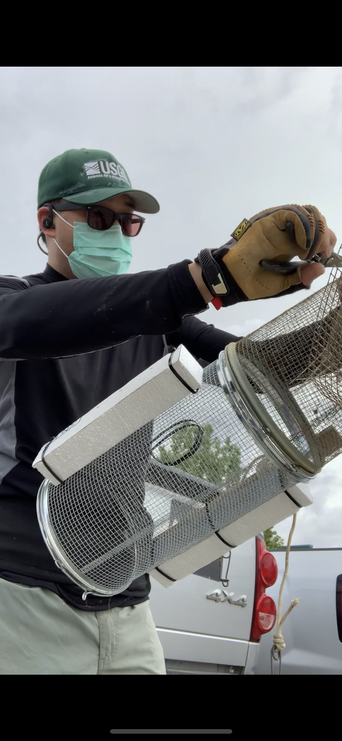 Man wearing a USGS hat and face mask holds up a cylindrical metal cage