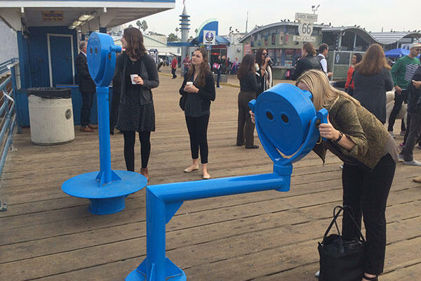 People stand on a pier near large viewers that look off the pier, one lady is looking through the viewer.