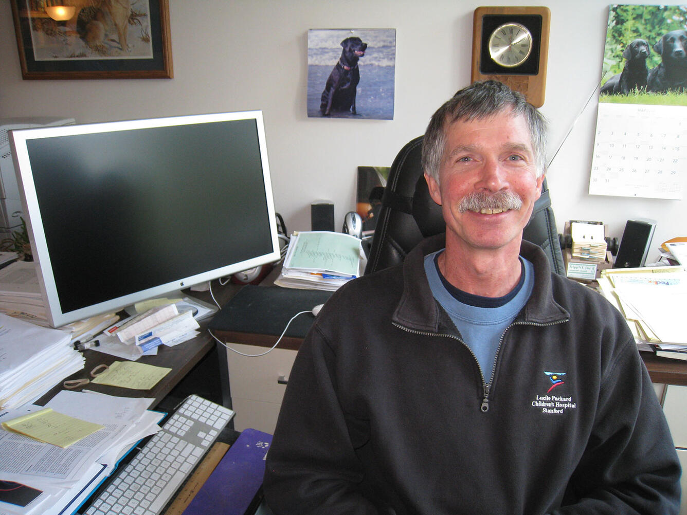 A smiling man in a blue t-shirt and black sweatshirt sits at a desk facing away from a computer screen