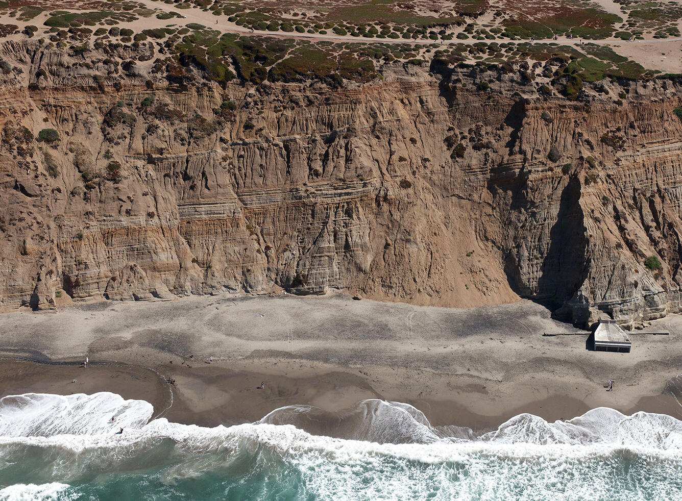 View looks at a high coastal cliff from out over the ocean, gentle waves on beach with historic, half-buried bunker.