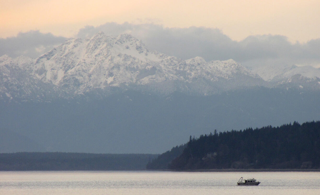 A small boat in a calm area of water with a spit of land in the distance, and snow-capped mountain peak in the far background.