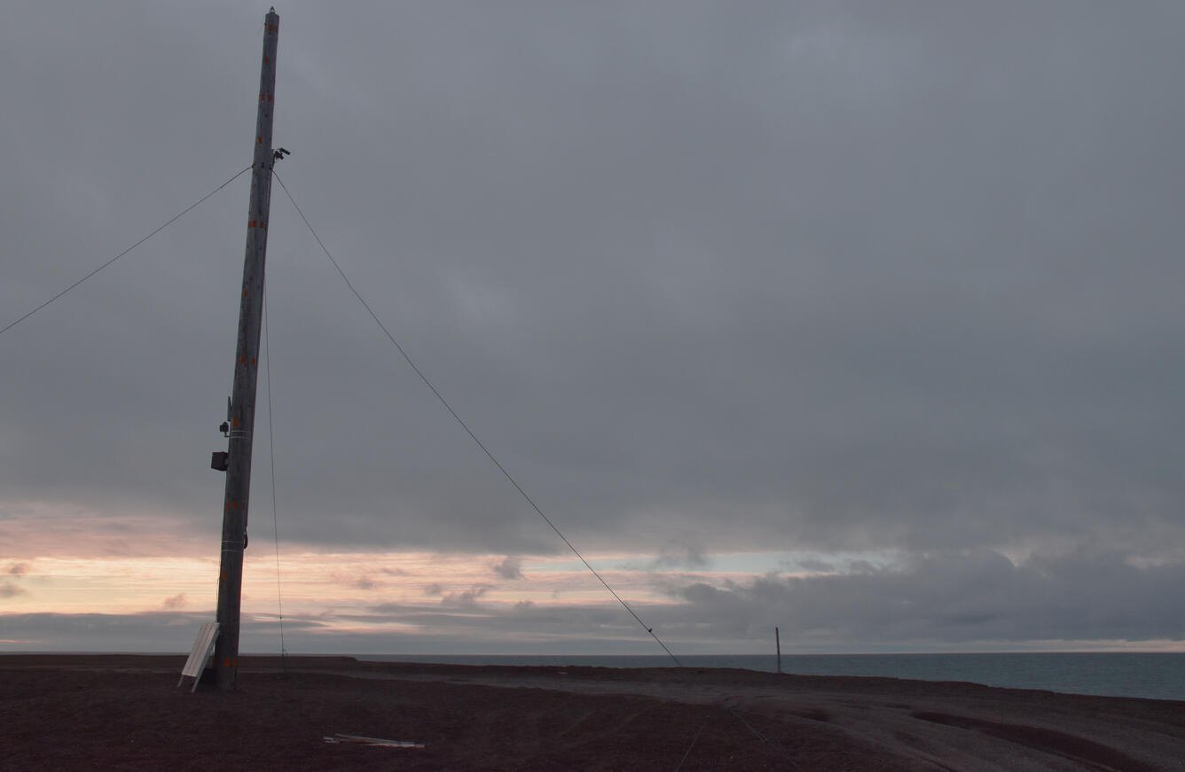 A pole with instruments strapped to it is supported by wires and stands on a coastal bluff with the ocean in the background.