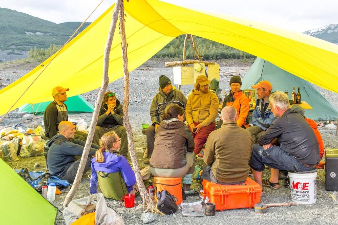 A group of people dressed warmly sit together under a tarp shelter listening to one man talk.