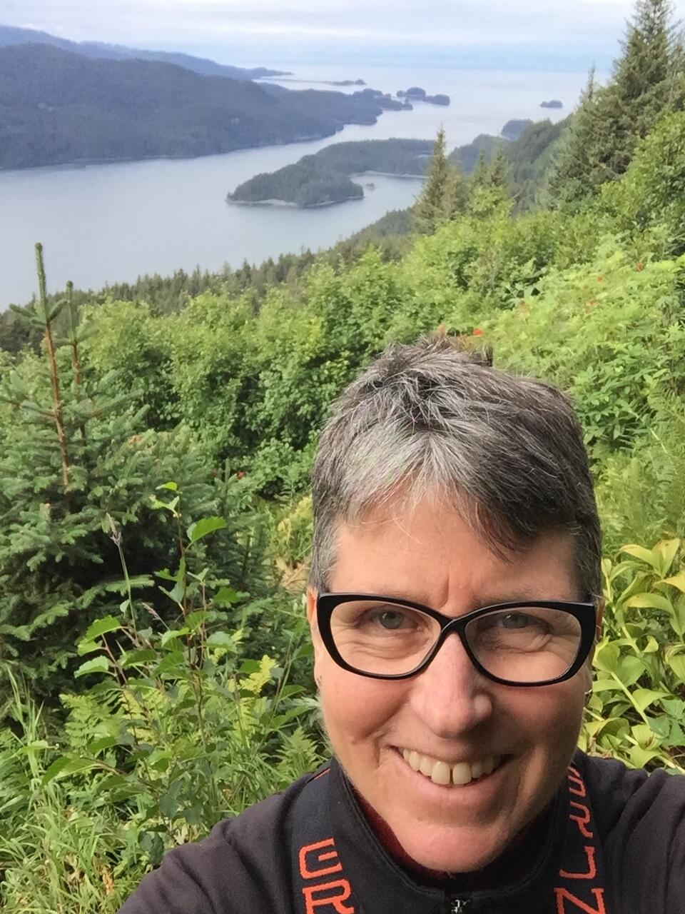 smiling female face with short hair and glasses, green shrubs and blue water in background