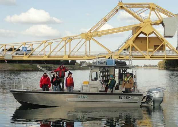 6 USGS crew members on a silver metal boat on a river with yellow bridge scaffolding in the background