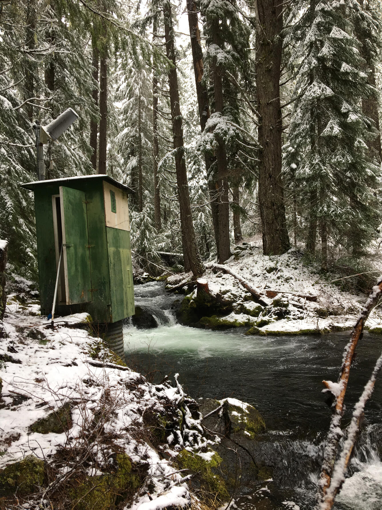 Photo of USGS gage house alongside a wooded stream during winter.