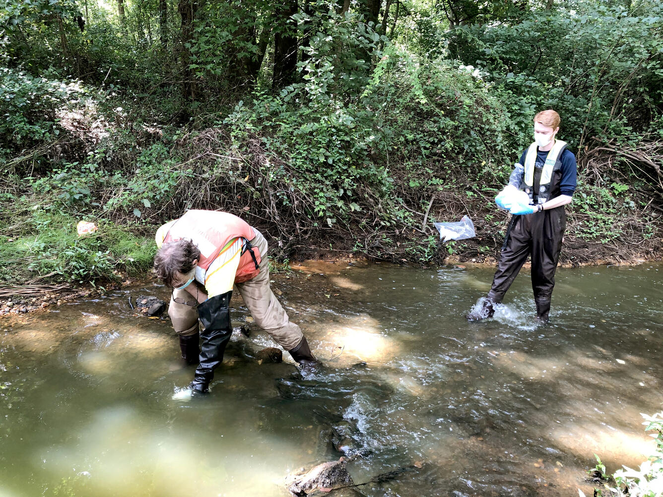 One scientist bends down to take a sample from a shallow stream while another scientist holds a sample container.