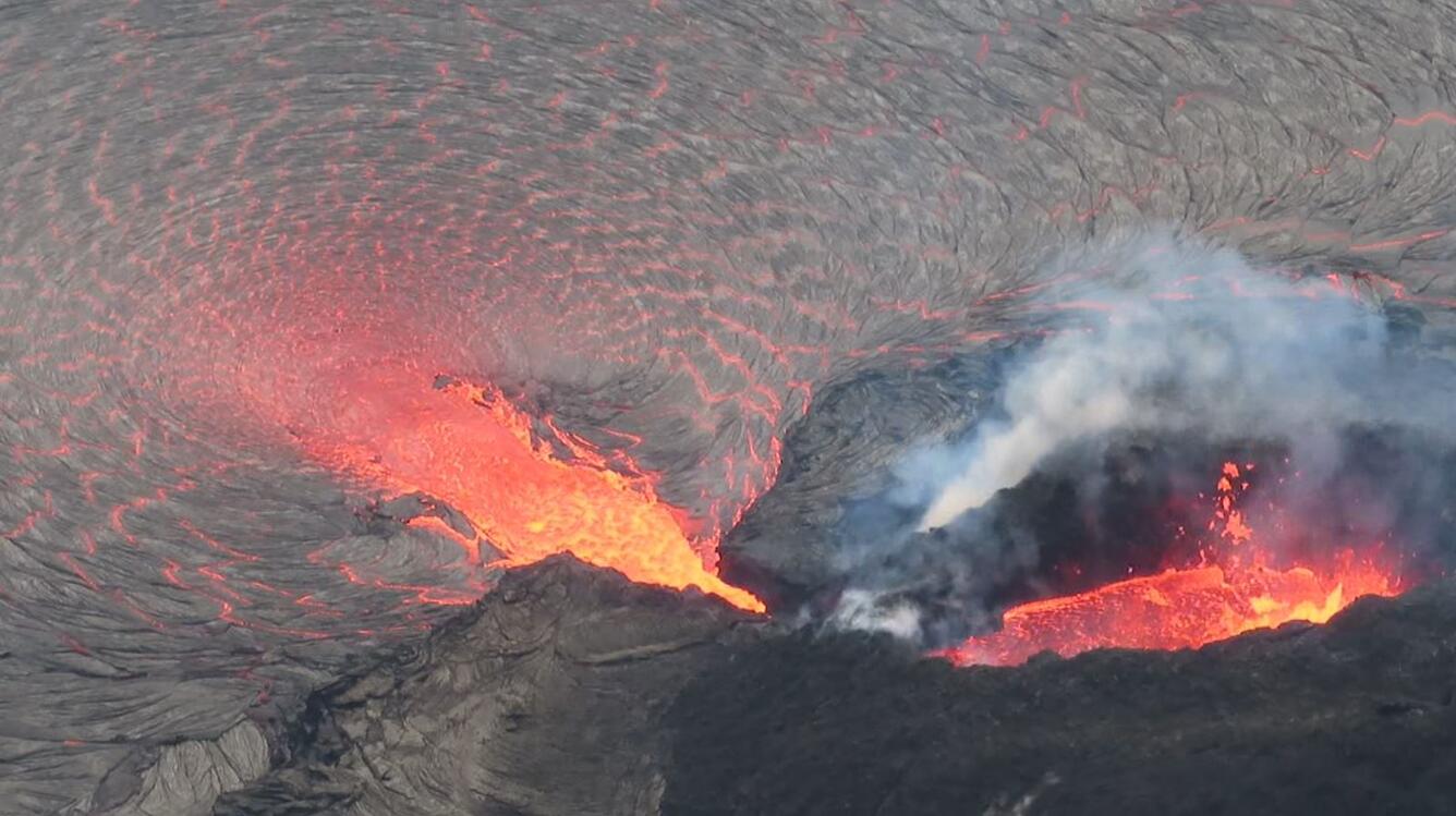 Telephoto color image of a small lava fountain within a darker spatter cone, which is supplying lava into a lava lake