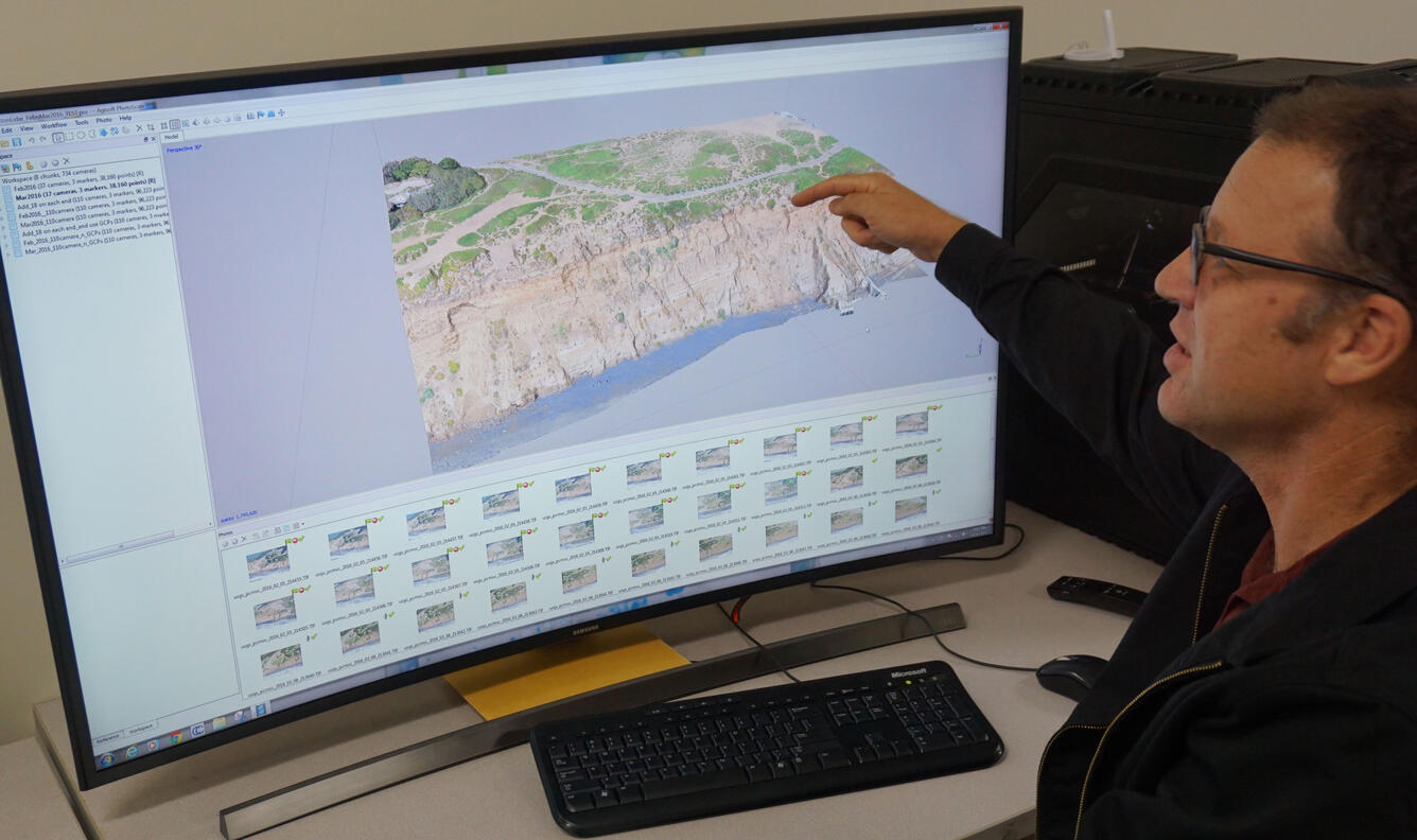 A man sits at a desk in front of a computer monitor and keyboard, pointing to the image and talking.