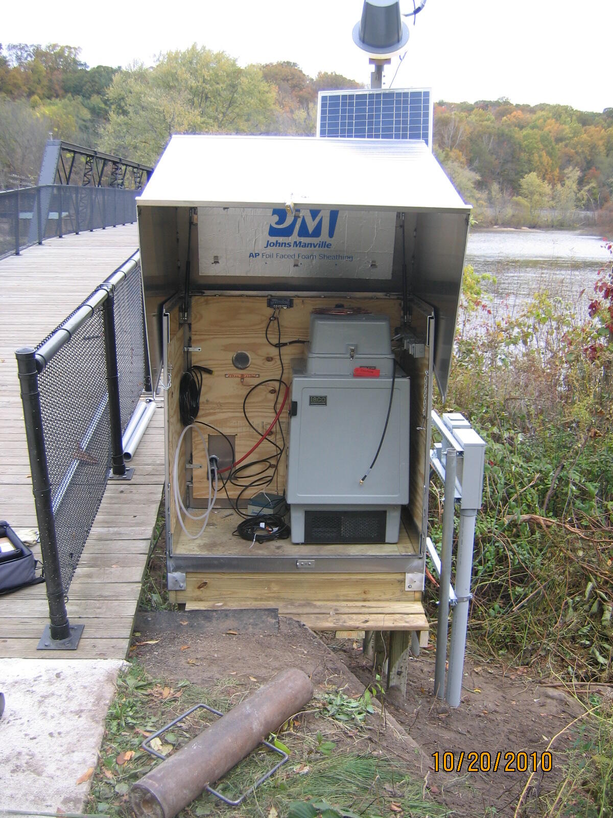 USGS Water-Quality equipment with cover opened at edge of river near a footbridge.
