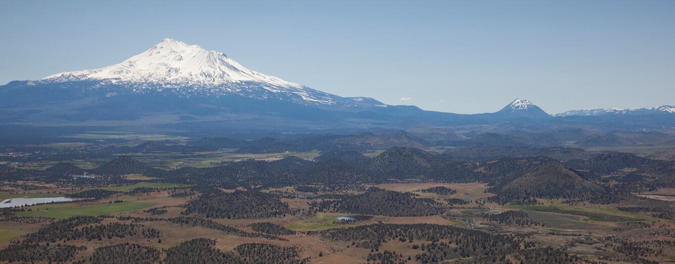 View of a snowcapped volcano with a green, hilly valley beneath and a small cinder cone volcano on the right.