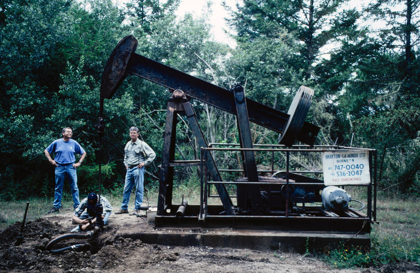 Two men stand by an oil well pumping unit, one man is crouched over the well, all in a forested location.