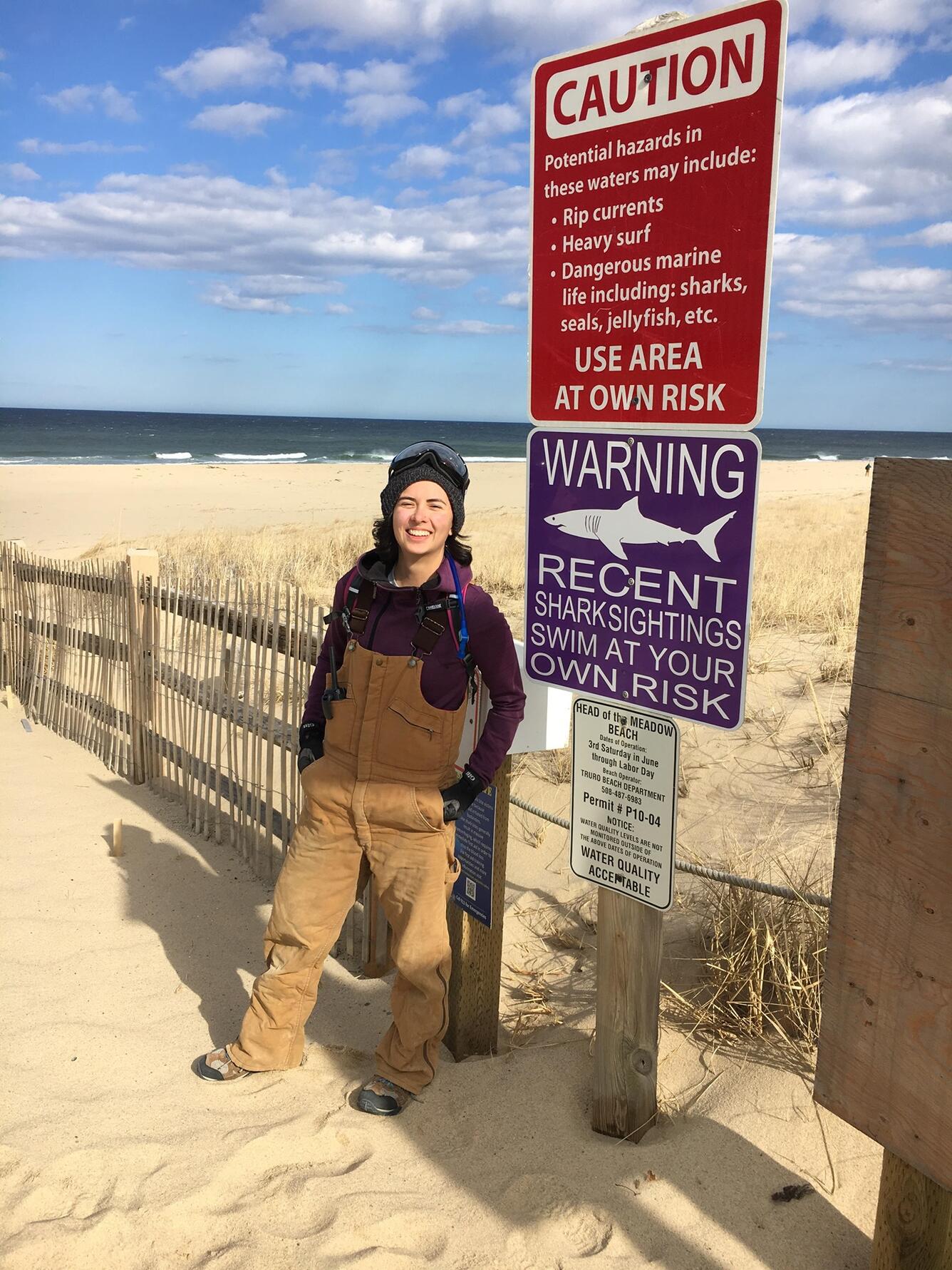 A woman stands on a sandy beach near a fence with the water in the background.