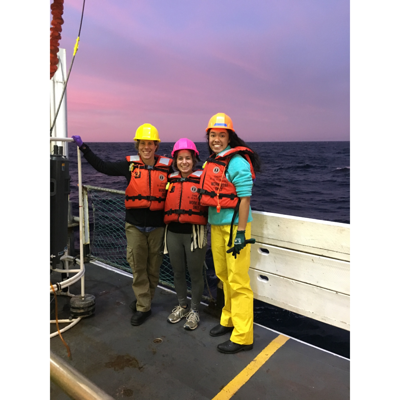 Three scientists stand on the deck of a ship in safety gear with the ocean and sunset in the background.