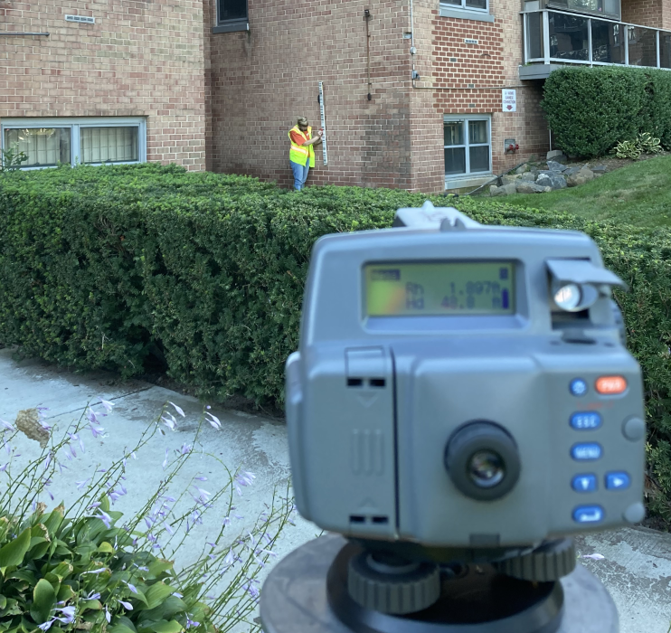 Image shows a survey theodolite facing a USGS scientist measuring a high-water mark against an apartment complex