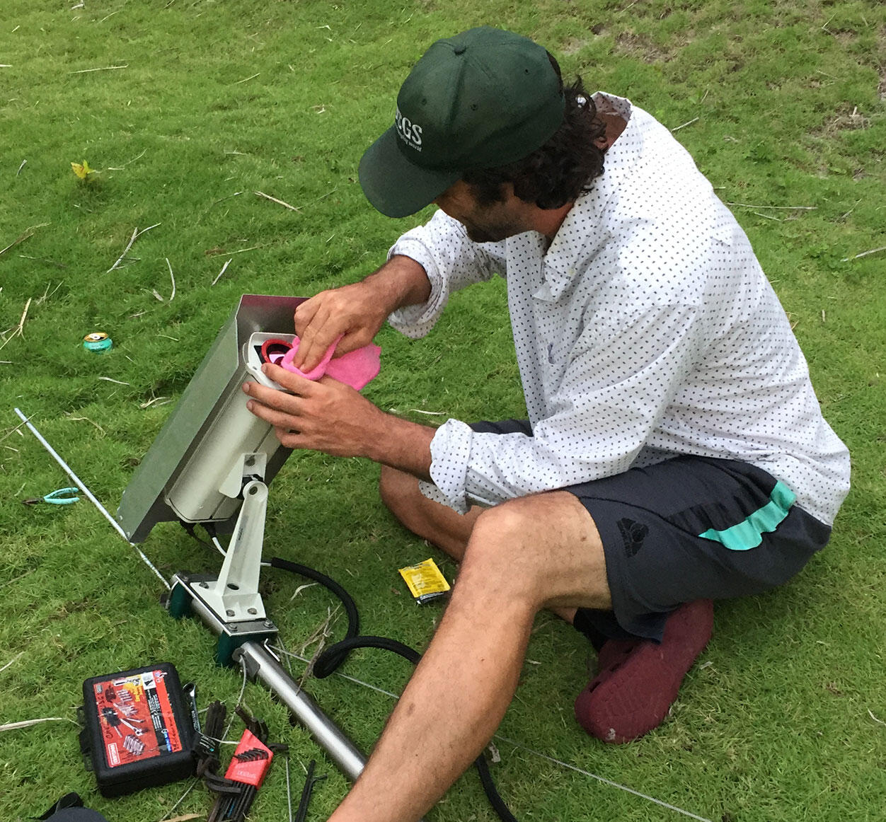 A man sitting on the grassy ground cleans a camera lens with a rag.