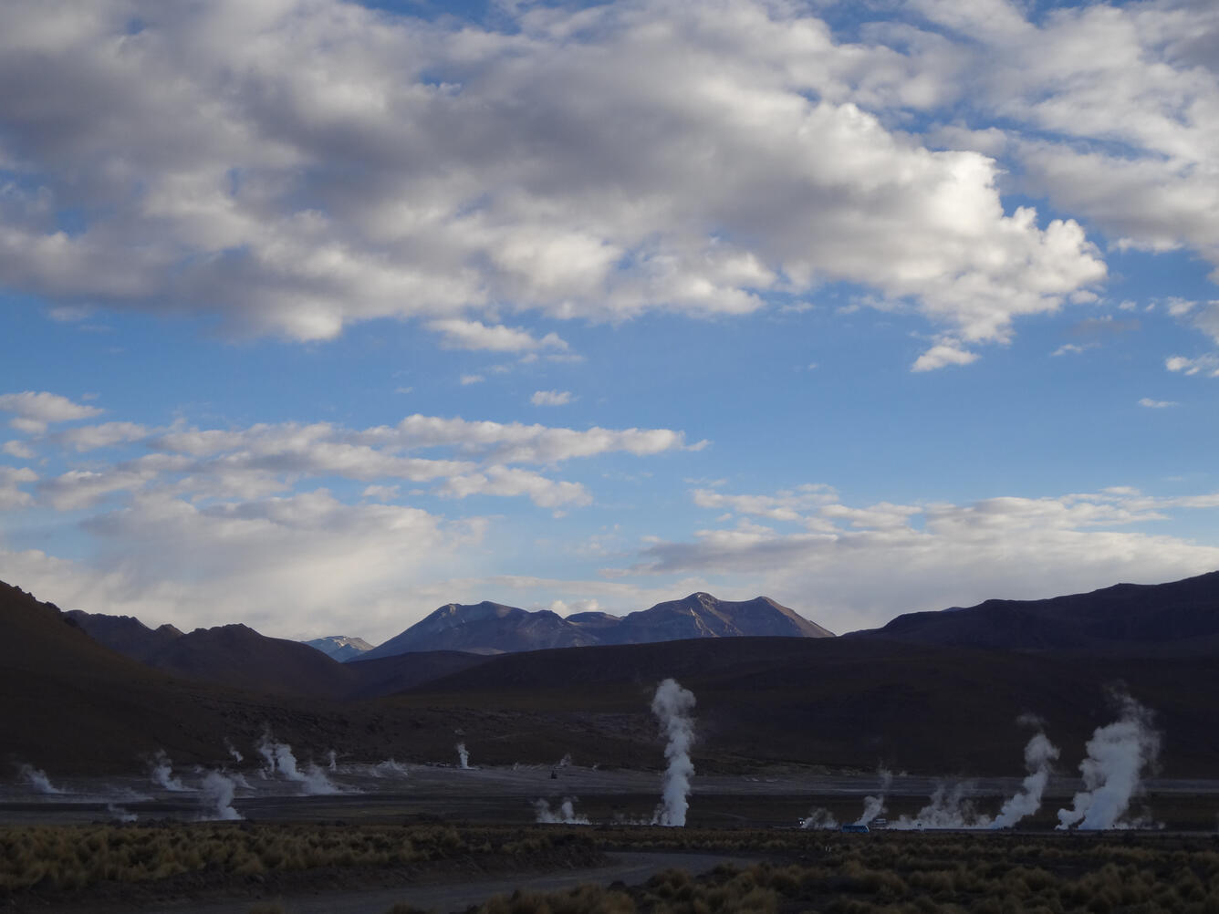 broad open valley, surrounded by mountains, with multiple wisps of steam coming out of the ground at several scattered locations