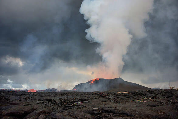 Far in the distance a hill spews smoke with lava pouring out, sky is dark with clouds, foreground is lifeless and burnt.