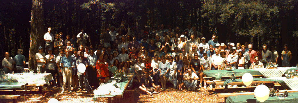 A large group of people stand posing for the camera at a picnic area under the trees with picnic tables in the foreground.