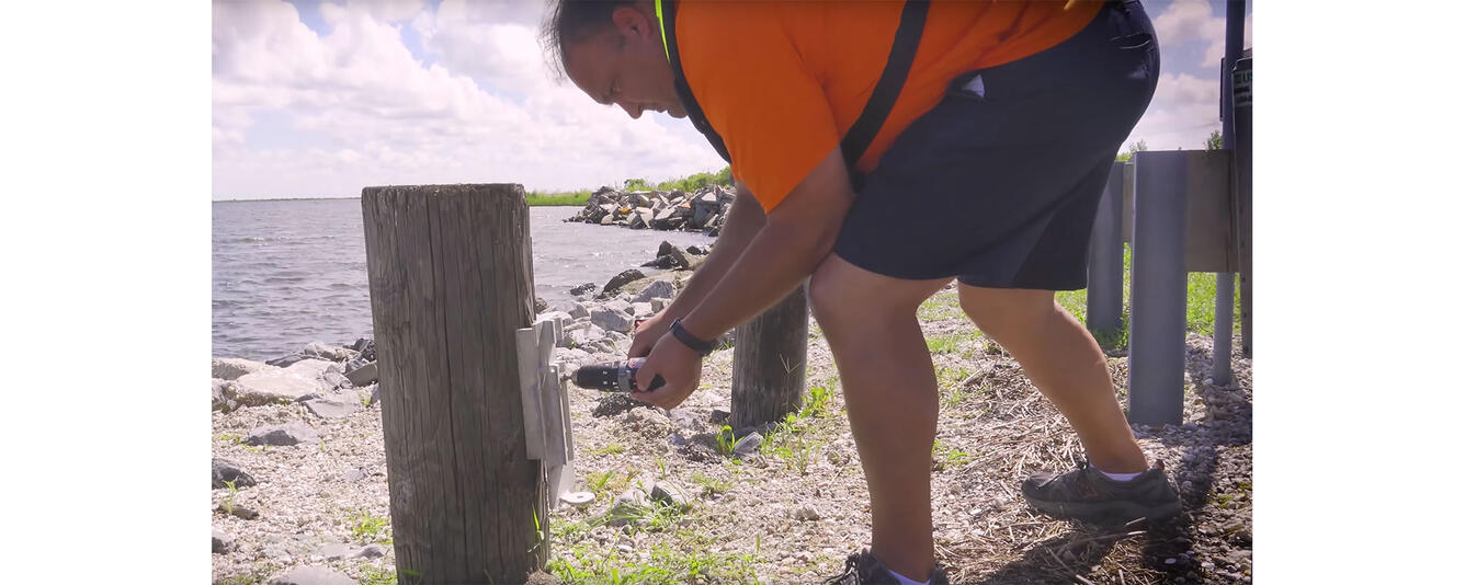 A man crouches next to a pole on a rocky, cobbly beach, and he is attaching an instrument bracket to the pole with a tool.