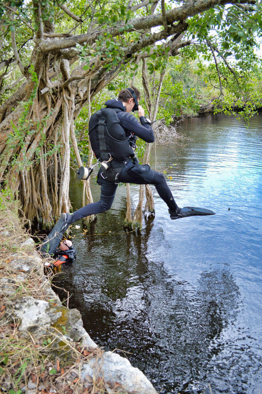 A diver jumps into water from a low, rocky cliff, and another diver is already in the water off to the side.