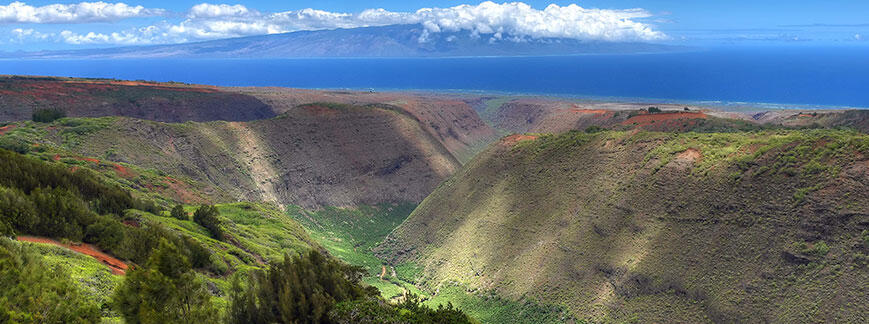 View from up high in mountains looking down into valleys with lush tropical foliage, an island can be seen way off in distance.