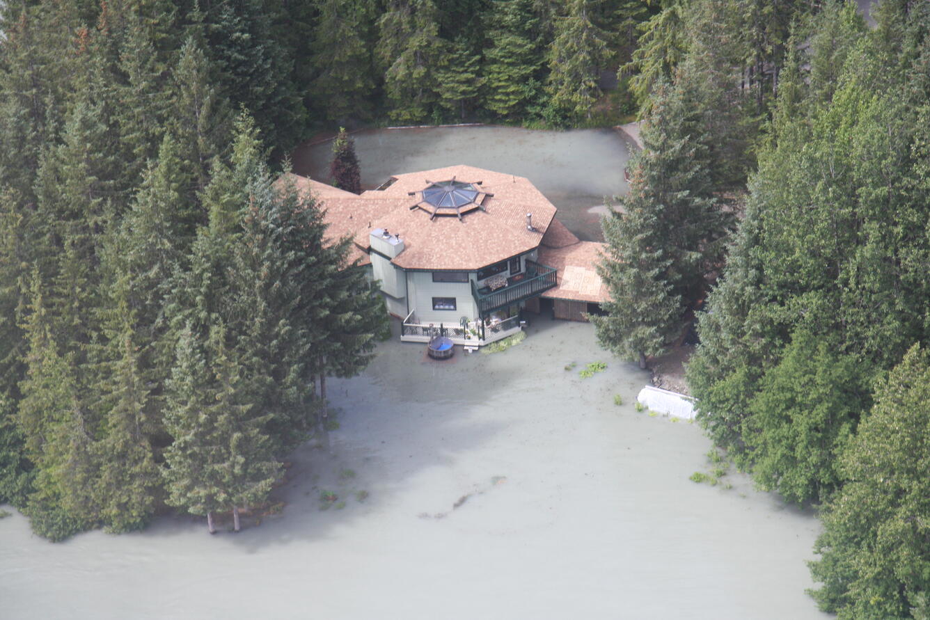 A home nestled between tall pine trees is surrounded by grey floodwaters reaching several feet up the first story walls. 