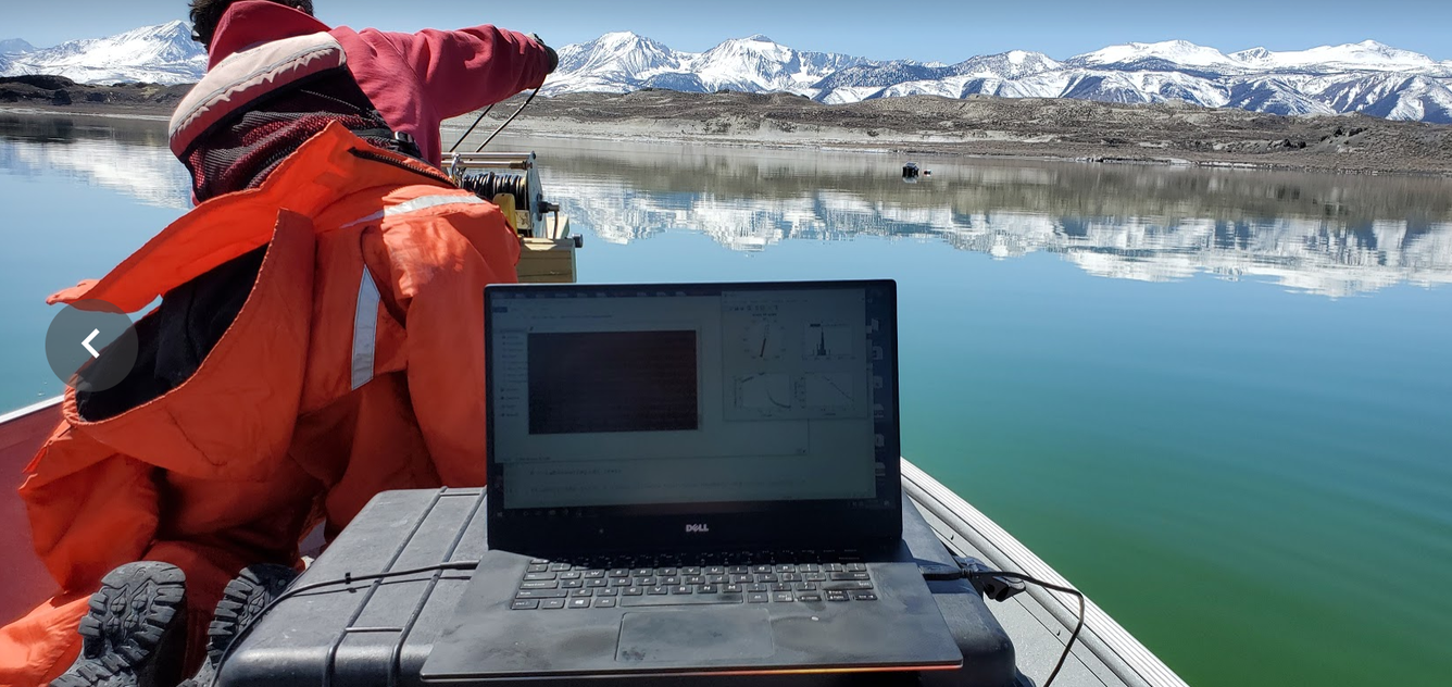 A red-coated scientist in a boat lowers a sensor to the bottom of a lake, while a laptop in the foreground shows temperatures.
