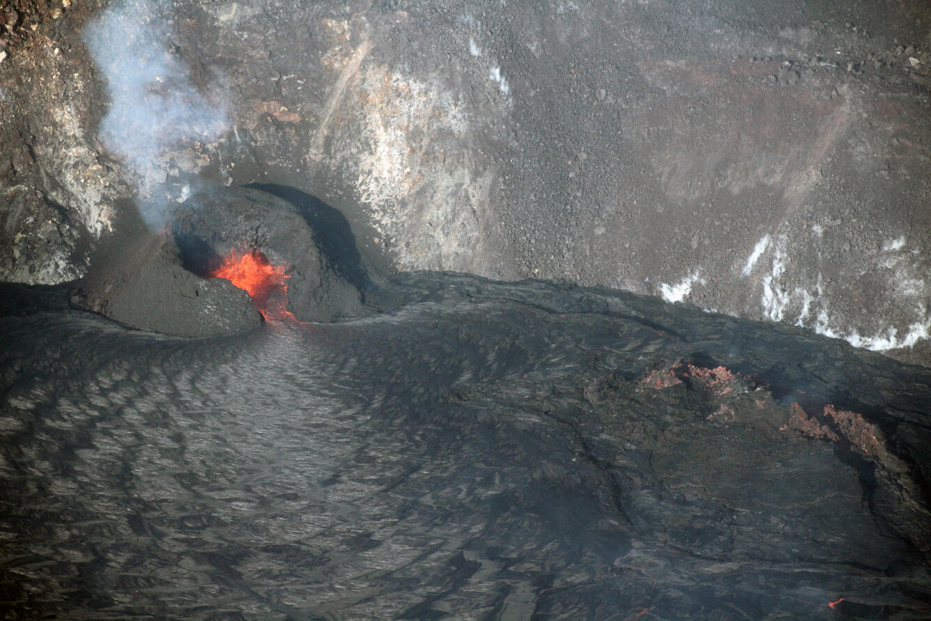 Color photo of a lava fountain within a black spatter cone at the edge of a lava lake